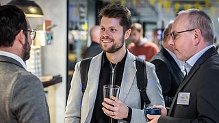 Foto, drei Männer in Businesskleidung unterhalten sich in einem modernen Innenraum, zwei halten Getränke in der Hand.