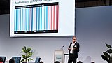 Foto, ein Redner hält einen Vortrag auf einer Bühne und zeigt Präsentationsfolien.