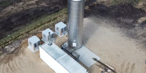 Foto, Luftaufnahme einer Wasserstoffanlage mit silbernem vertikalem Tank, weißem rechteckigem Gebäude und drei Personen am Eingang