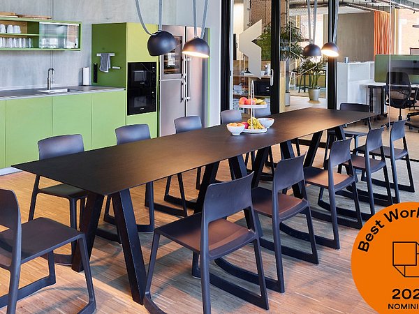 Foto, Moderner Besprechungsraum mit langem schwarzem Tisch, mehreren schwarzen Stühlen, grüner Küchenzeile im Hintergrund und großen Fenstern zu einem angrenzenden Bürobereich.