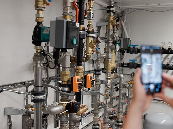 Mehrere vertikale Metallrohre mit Messgeräten und Ventilen in einem Technikraum, im Vordergrund zwei Personen, eine hält ein Smartphone zum Fotografieren.