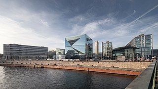 Foto, Panoramasicht, rechts befindet sich der Berliner Hauptbahnhof, in der Mitte ein großes quaderförmiges, verspiegeltes Gebäude, links weitere Hochhäuser. Im Vordergrund verläuft die Spree.