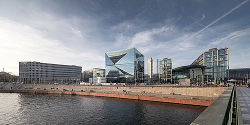 Foto, Panoramasicht, rechts befindet sich der Berliner Hauptbahnhof, in der Mitte ein großes quaderförmiges, verspiegeltes Gebäude, links weitere Hochhäuser. Im Vordergrund verläuft die Spree.