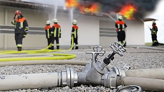 Foto, im Vordergrund auf dem Boden liegend ein Feuerwehrschlauch mit Sperrventil, im Hintergrund einige Feuerwehrmitarbeiter beim Löschen einer Photvolatik-Anlage auf dem Dach eines Gebäudes.