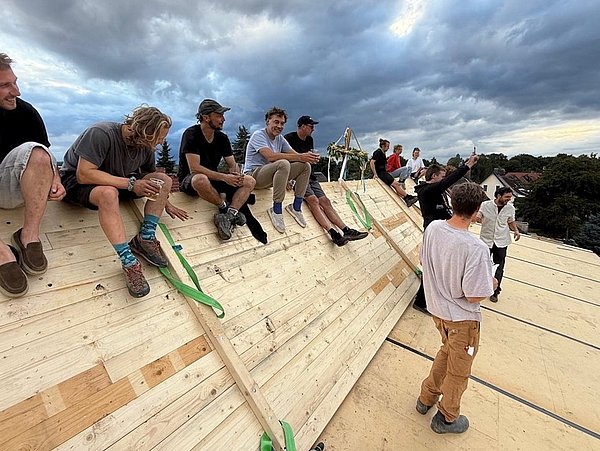 Foto, Mehrere Personen sitzen auf einer schrägen Holzfläche mit grünen Gurten, während andere davor stehen und sich unterhalten