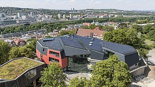 Foto, Luftaufnahme eines modernen Gebäudes mit schwarzem Dach und roten Wänden, umgeben von Bäumen, mit Stadt und Hügeln im Hintergrund