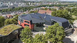 Foto, Luftaufnahme eines modernen Gebäudes mit schwarzem Dach und roten Wänden, umgeben von Bäumen, mit Stadt und Hügeln im Hintergrund