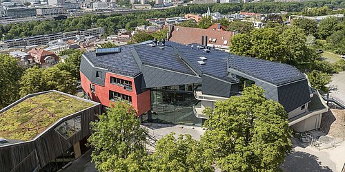 Foto, Luftaufnahme eines modernen Gebäudes mit schwarzem Dach und roten Wänden, umgeben von Bäumen, mit Stadt und Hügeln im Hintergrund