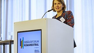 Foto, eine Frau steht auf einem Podium und hält eine Rede, neben ihr stehen Wassergläser.