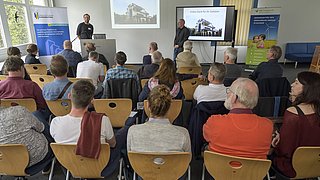 Foto, zwei Männer halten einen Vortrag vor sitzendem Publikum in einem hellen Raum mit Fenstern und Präsentationsbildschirm.