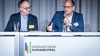 Foto, zwei Männer in Anzügen sitzen an einem Tisch mit Mikrofonen und Wassergläsern, vor einem Banner mit dem Schriftzug "Gebäudeforum Klimaneutral".