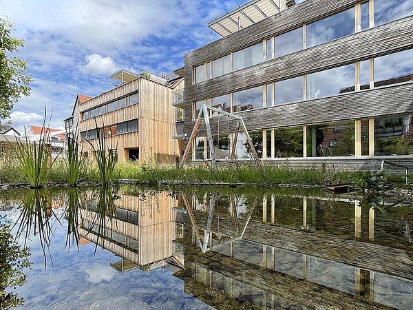 Foto, Außenansicht zweier mehrstöckiger Gebäude mit Holzfassaden. Im Vordergrund befindet sich ein Teich.