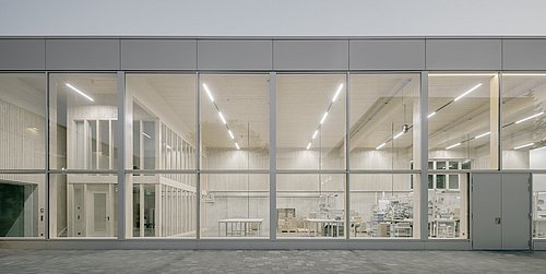 Foto, Fassade eines modernen Gebäudes mit großen Glasfenstern, durch die ein heller, minimalistisch eingerichteter Innenraum mit Tischen und Regalen sichtbar ist.