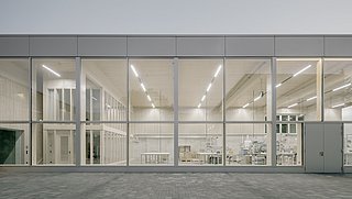 Foto, Fassade eines modernen Gebäudes mit großen Glasfenstern, durch die ein heller, minimalistisch eingerichteter Innenraum mit Tischen und Regalen sichtbar ist.