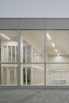 Foto, Fassade eines modernen Gebäudes mit großen Glasfenstern, durch die ein heller, minimalistisch eingerichteter Innenraum mit Tischen und Regalen sichtbar ist.