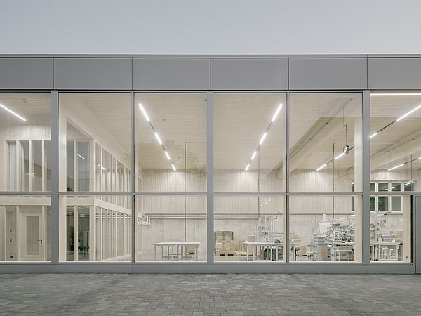 Foto, Fassade eines modernen Gebäudes mit großen Glasfenstern, durch die ein heller, minimalistisch eingerichteter Innenraum mit Tischen und Regalen sichtbar ist.