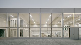 Foto, Fassade eines modernen Gebäudes mit großen Glasfenstern, durch die ein heller, minimalistisch eingerichteter Innenraum mit Tischen und Regalen sichtbar ist.