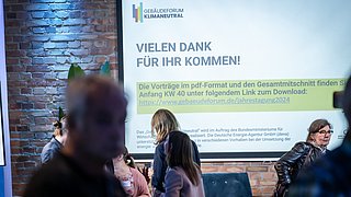 Foto, Leinwand mit Text 'Vielen Dank für Ihr Kommen!' und Link zur Jahrestagung 2024 des Gebäudeforum Klimaneutral, Menschen im Gespräch vor der Leinwand