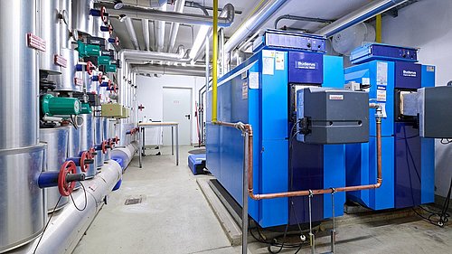 Foto, Technikraum mit blauen Heizkesseln, Rohrleitungen und Ventilen an der Wand.