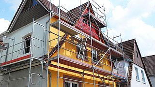 Foto, Haus mit gelber Fassade und braunem Dach mit Gerüst für Bauarbeiten.