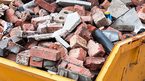 Foto, gelber Container gefüllt mit zerbrochenen Ziegelsteinen und Bauschutt.