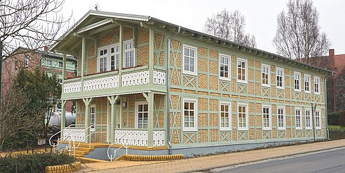 Foto, Zweistöckiges Fachwerkhaus mit gelben Ziegeln und grünen Holzbalken an einer Straße