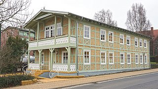Foto, Zweistöckiges Fachwerkhaus mit gelben Ziegeln und grünen Holzbalken an einer Straße