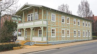 Foto, Zweistöckiges Fachwerkhaus mit gelben Ziegeln und grünen Holzbalken an einer Straße