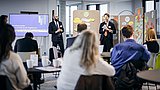 Foto, Zwei Personen in Anzügen präsentieren vor Pinnwänden mit bunten Notizzetteln, Publikum sitzt an Tischen in modernem Konferenzraum.
