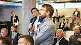 Foto, ein Redner hält ein Mikrofon in der Hand und wendet sich in einer modernen Konferenzumgebung an das Publikum. Die Zuhörer hören im Hintergrund aufmerksam zu.