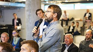 Foto, ein Redner hält ein Mikrofon in der Hand und wendet sich in einer modernen Konferenzumgebung an das Publikum. Die Zuhörer hören im Hintergrund aufmerksam zu.