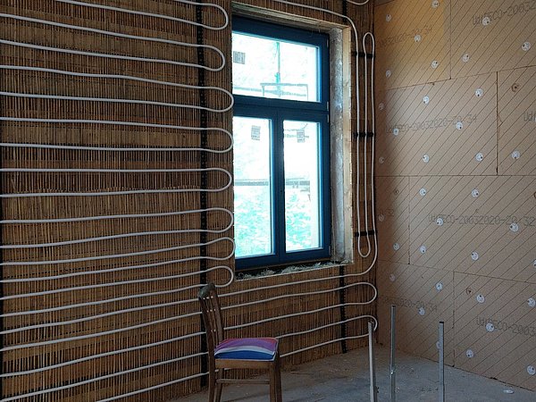 Foto, Raum mit Holzvertäfelung, in dem weiße Heizrohre an der Wand und auf dem Boden verlegt sind, vor einem Fenster mit schwarzem Rahmen steht ein Stuhl mit blau-rotem Sitzkissen