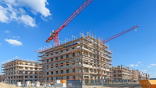 Foto, mehrere mehrstöckige Rohbauten auf einer Baustelle mit einem großen Baukran vor blauem Himmel mit Wolken.