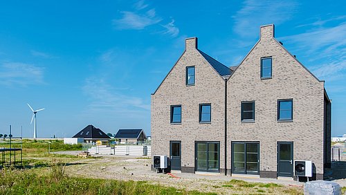 Foto, Moderne zweistöckige Wohnhäuser unter einem strahlend blauen Himmel, mit einer Windkraftanlage im Hintergrund und Baumaterialien in der Nähe.