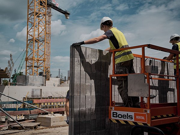 Foto, zwei Bauarbeiter in Sicherheitswesten und Helmen auf einer Arbeitsbühne, die Betonsteine positionieren, im Hintergrund ein Kran und eine Baustelle.