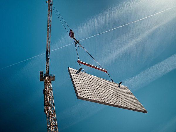 Foto, gelber Baukran hebt eine große Mauerwand vor blauem Himmel mit Kondensstreifen.