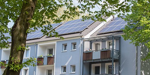 Foto, Mehrstöckiges Wohngebäude mit blauer Fassade, Balkonen und Solarpaneelen auf dem Dach, umgeben von grünen Bäumen