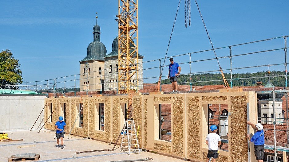 Foto, Baustelle eines Gebäudes dessen Wände aus Stroh gefertigt sind. Im Hintergrund stehen zwei Kirchtürme.