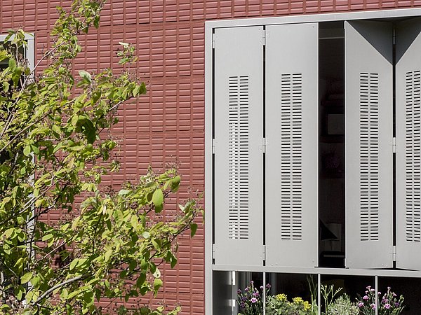 Graue Fensterläden mit vertikalen Lüftungsschlitzen, geöffnet vor einer roten Wand mit quadratischem Muster. Im Vordergrund ein grüner Baum.