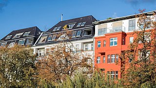 Foto, Mehrfamilienhäuser mit dunklem Dach und einem orangefarbenen Gebäude im Vordergrund, umgeben von Bäumen und blauem Himmel.