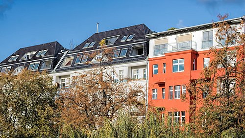 Foto, Mehrfamilienhäuser mit dunklem Dach und einem orangefarbenen Gebäude im Vordergrund, umgeben von Bäumen und blauem Himmel.