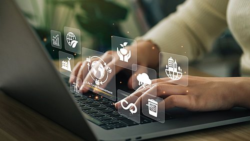 Foto, Nahaufnahme einer Person, die an einem Schreibtisch sitzend an einem Laptop arbeitet; über dem Laptop schweben digital hinzugefügte Symbole zum Thema Lebenszyklus. 