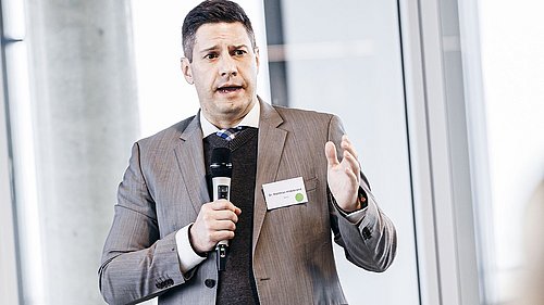 Foto, Mann in grauem Anzug und schwarzem Pullover hält Mikrofon und gestikuliert mit der freien Hand.