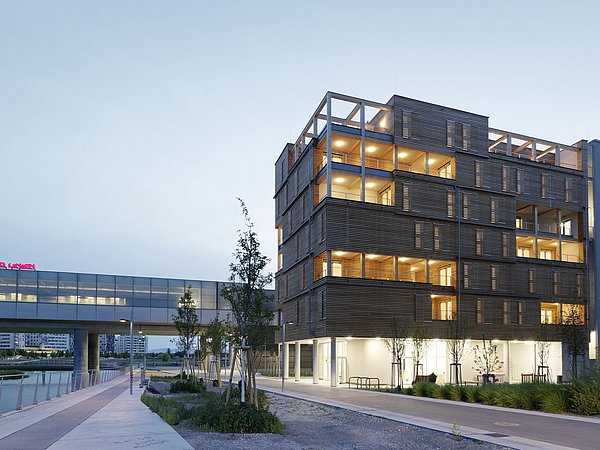 Foto, Modernes sechsstöckiges Gebäude mit Holzfassade und beleuchteten Balkonen am Wasserufer bei Dämmerung