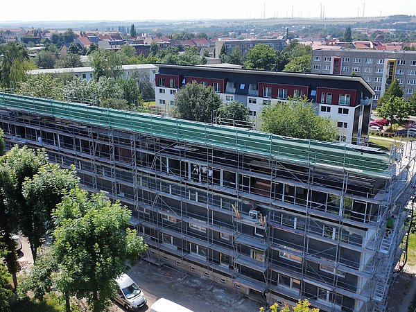 Mehrstöckiges Wohngebäude mit Gerüst, umgeben von Bäumen und weiteren Wohnhäusern des gleichen Bautypus im Hintergrund.