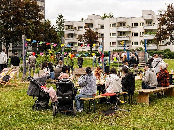 Menschen unterschiedlichen Alters sitzen und stehen auf einer Wiese in einem Wohnquartier, umgeben von Wohngebäuden, mit bunten Wimpeln und Kinderwagen, beim gemeinschaftlichen Beisammensein