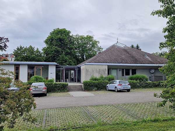 Foto, Flaches, modernes Kirchengebäude mit Kreuz auf dem Dach, umgeben von Bäumen und Parkplätzen mit zwei geparkten Autos