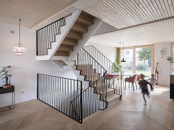 Foto, Offenes Wohnzimmer mit moderner Holztreppe, zwei spielende Kinder, großer Fensterfront und Essbereich mit rosa Stühlen