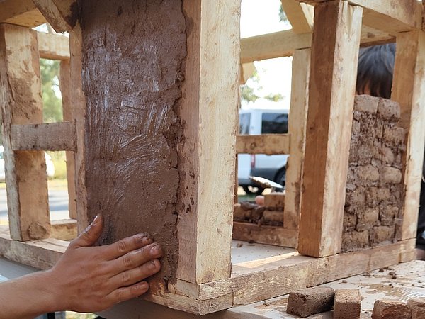 Grundschulkinder bauen bei Projekttag Naturbau Campinis ein Fachwerkhaus aus Holzrahmen und Lehm. Zwei Hände formen Lehm an der Wand des Miniatur-Fachwerkhaus-Modells.
