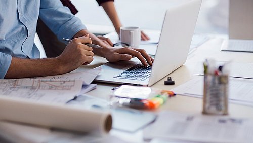 Foto, Eine Person mit einem blauen Hemd tippt auf einem Laptop. Auf dem Tisch liegen Pläne, Dokumente und Schreibutensilien. Eine Tasse steht neben dem Laptop.